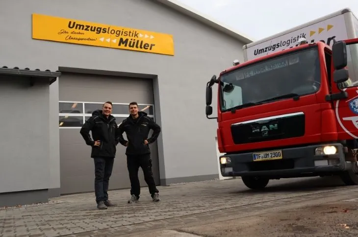 zwei umzugshelfer stehen vor firmengebaeude mit umzugswagen